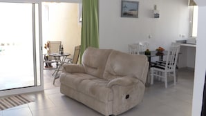 Ein Wohnzimmer mit beigefarbenem Sofa, ein Essbereich mit Tisch und Stühlen sowie eine Küchenzeile mit Arbeitsfläche und Stühlen.