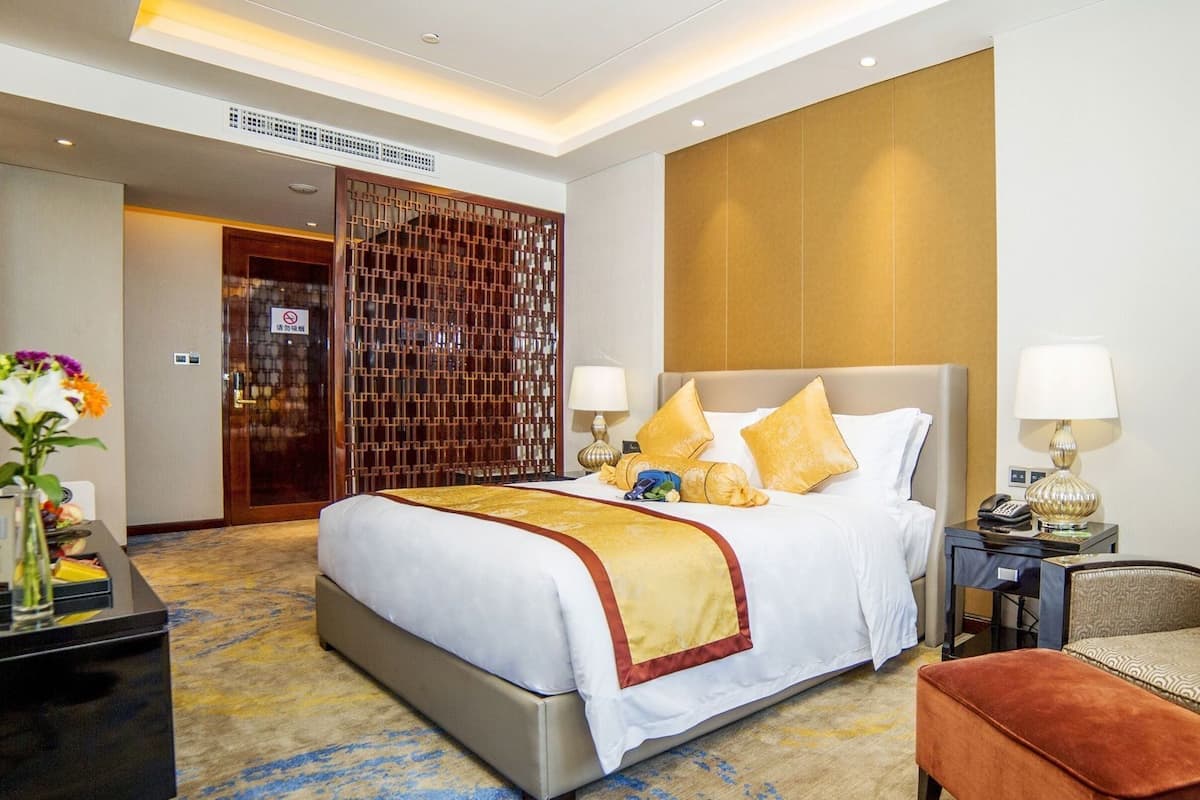 Quarto de hotel com cama grande, mesas de cabeceira, uma cadeira e escrivaninha com luminária.