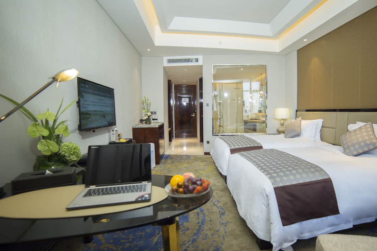 Quarto de hotel com escrivaninha, laptop, cesta de frutas, duas camas e banheiro visível por uma porta de vidro.