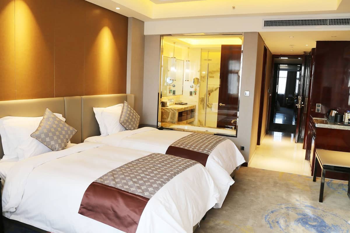 Quarto de hotel com duas camas, um espelho grande, uma escrivaninha e um banheiro visível através de uma porta de vidro.