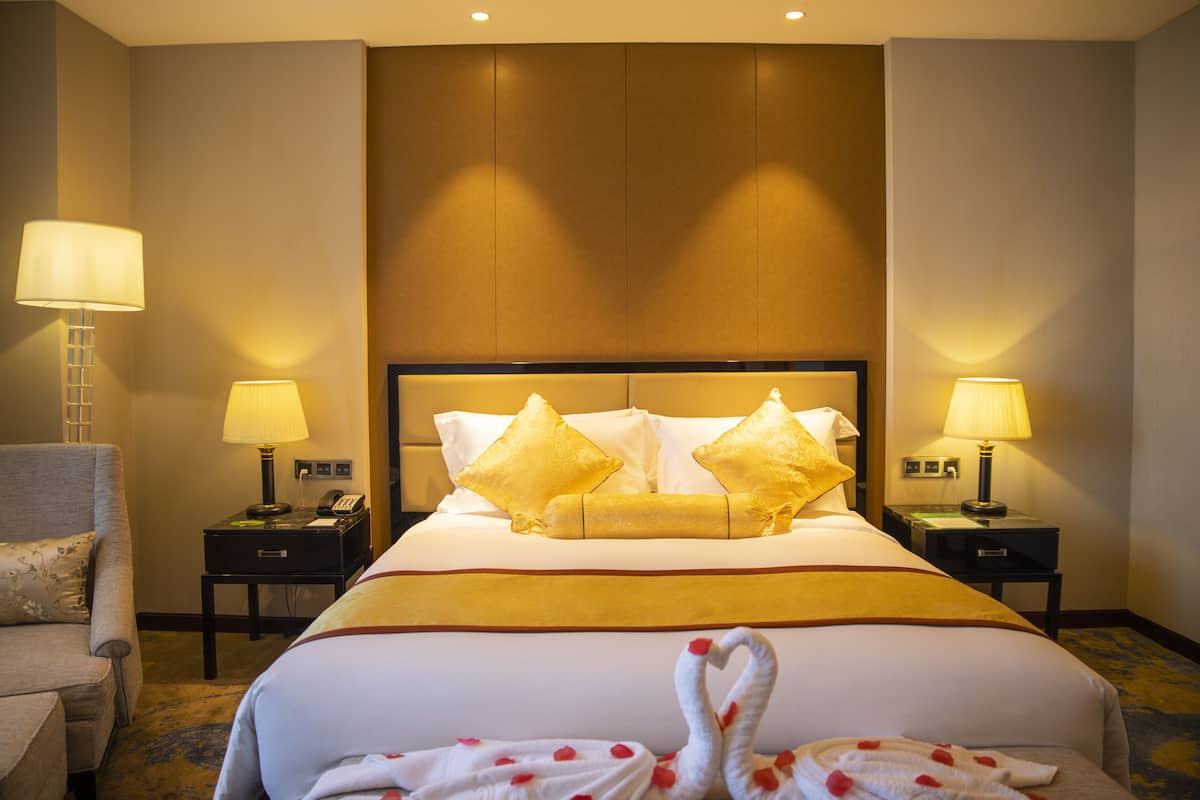 Quarto de hotel com uma cama grande, duas mesas de cabeceira com abajures, uma cadeira e uma toalha em formato de cisne decorativa sobre a cama.