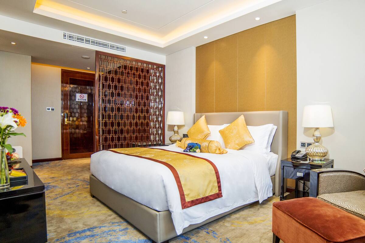 Quarto de hotel com uma cama grande, duas mesas de cabeceira, uma cadeira e um painel decorativo na parede.