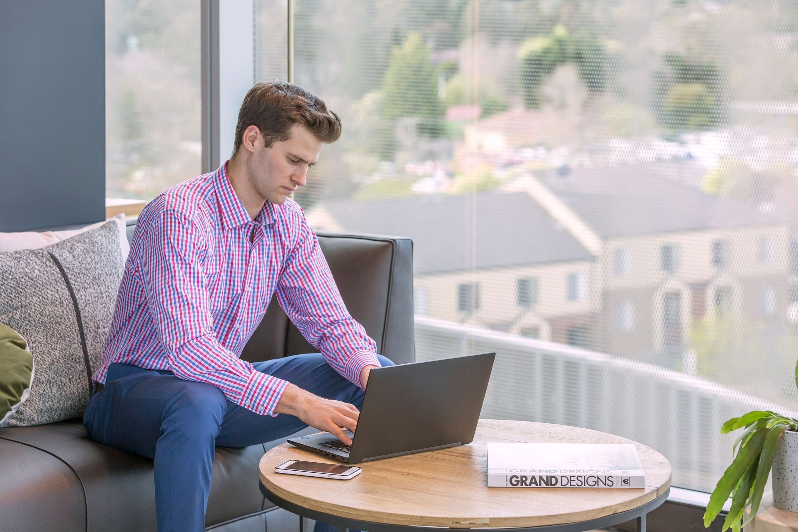 Iemand zit op een bank en gebruikt een laptop, met een boek getiteld 'GRAND DESIGNS' op een klein tafeltje ernaast.