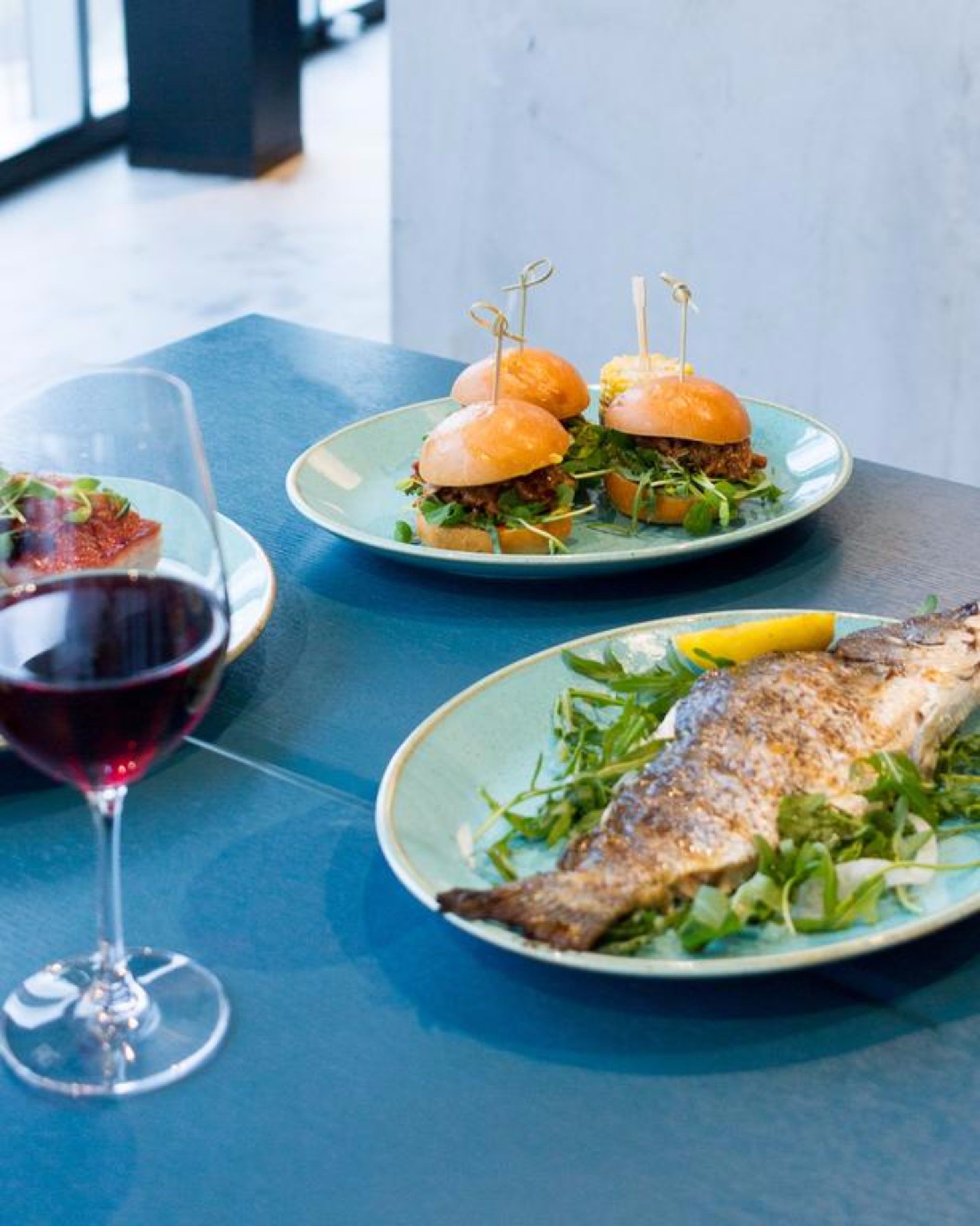 A table with a plate of fish, two burgers, and a glass of red wine.