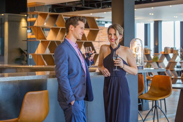 Un uomo e una donna sono in piedi al bar in un'accogliente hall d'hotel moderna, con scaffalature geometriche e zone salotto.