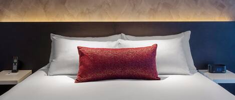 A neatly made bed with white and red pillows, a bedside table with a phone and a clock, and a textured wall behind the bed.