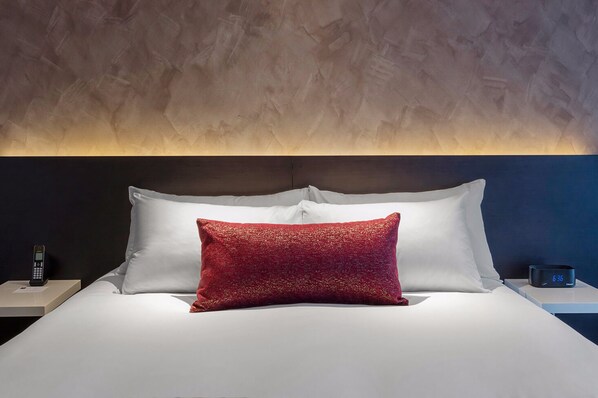Una cama bien hecha con almohadas blancas y rojas, una mesita de noche con un teléfono y un reloj, y una pared con textura detrás de la cama.