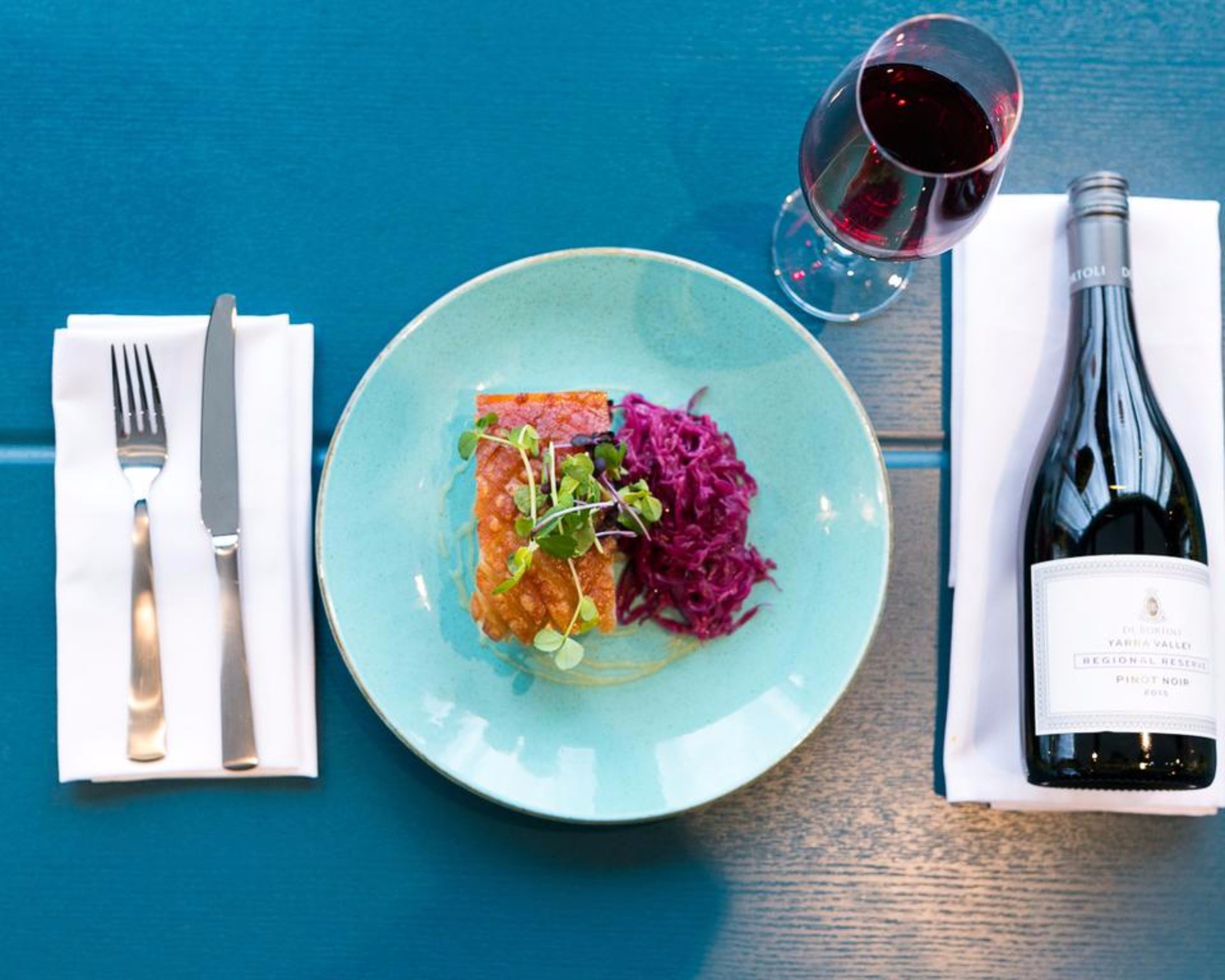 Prato de comida com uma garrafa de vinho e talheres sobre uma mesa azul.