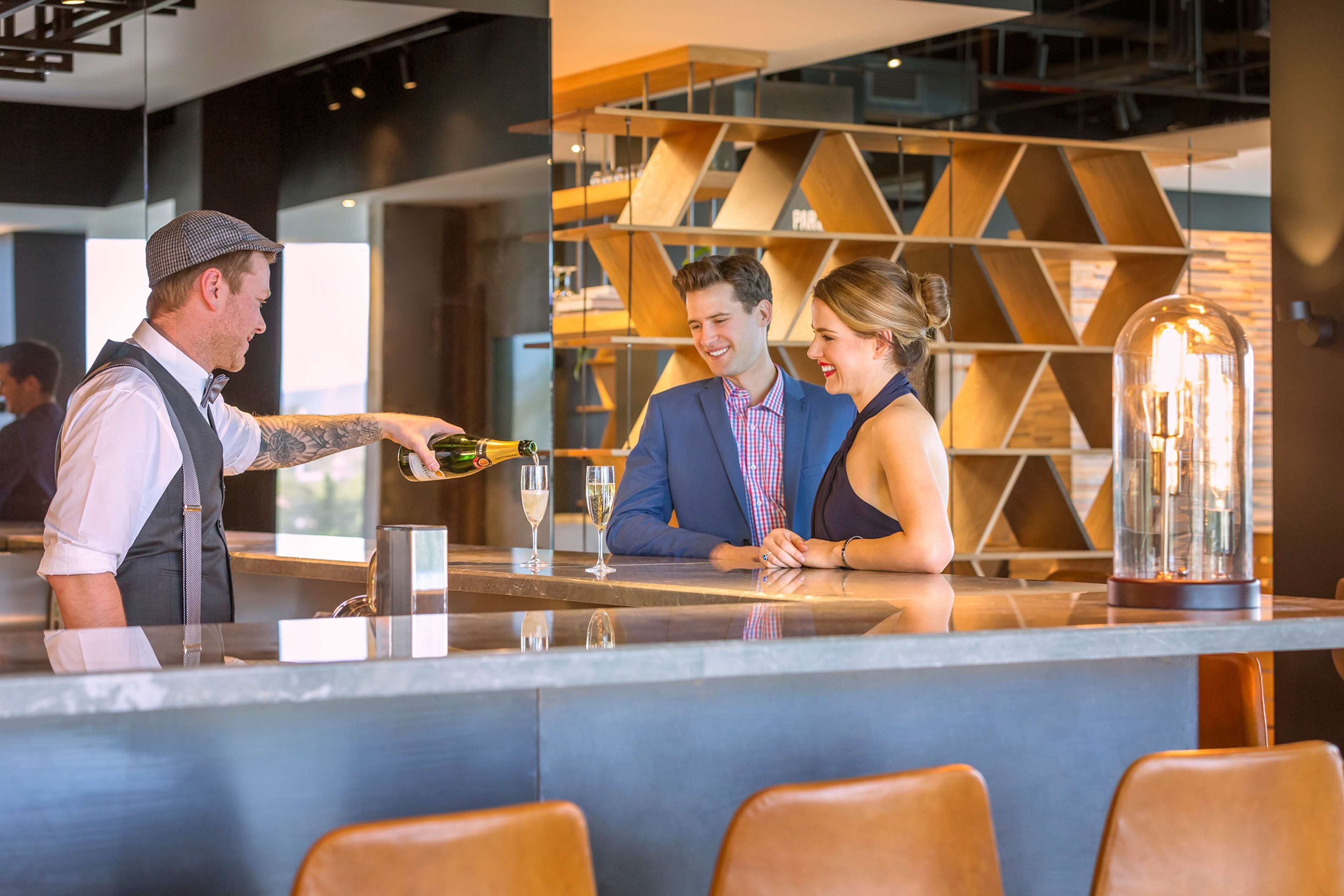 Zona de bar con un barman sirviendo una bebida, una pareja sentada en una mesa y una decoración interior moderna con estanterías de madera y una estructura de vidrio.