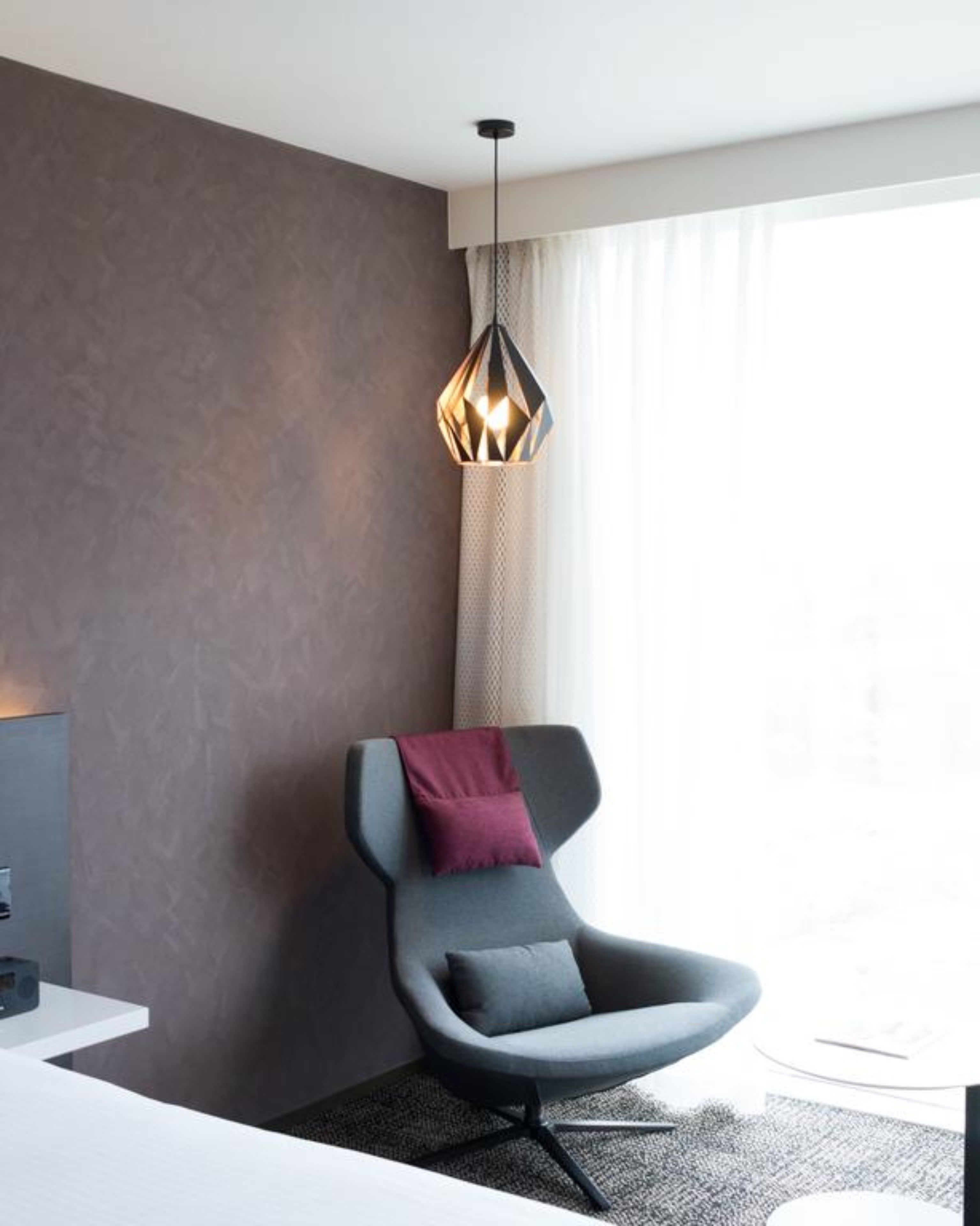 A modern room with a grey chair, a pendant light, and a large window with curtains.