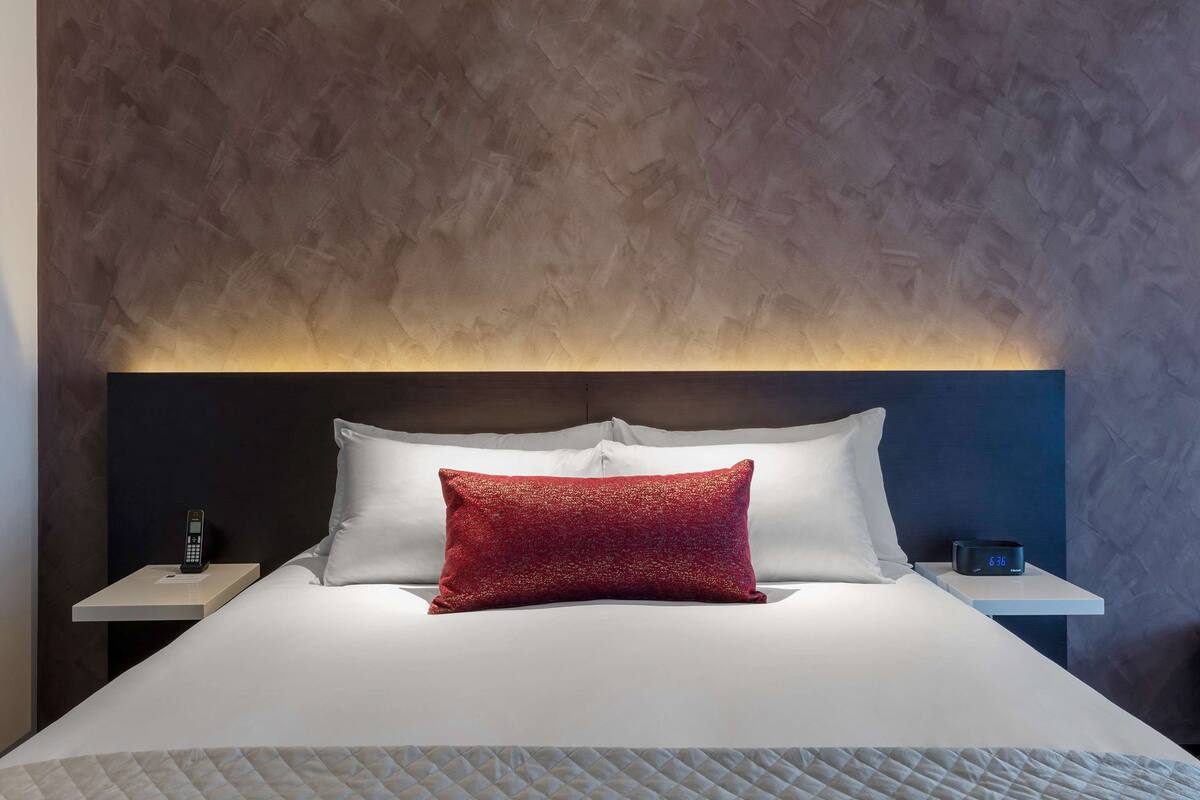 A neatly made bed with white linens and a red accent pillow, a bedside table with a phone and a digital clock, and a textured wall behind the bed.