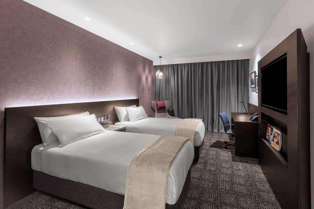 A hotel room with two beds, a desk, a TV, and a large window with curtains.