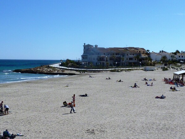 Une plage avec un grand bâtiment en arrière-plan, quelques personnes sur le sable et l’océan.
