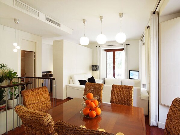 Una sala de estar moderna con una mesa de comedor de madera, sillas de mimbre, un sofá blanco y un televisor.