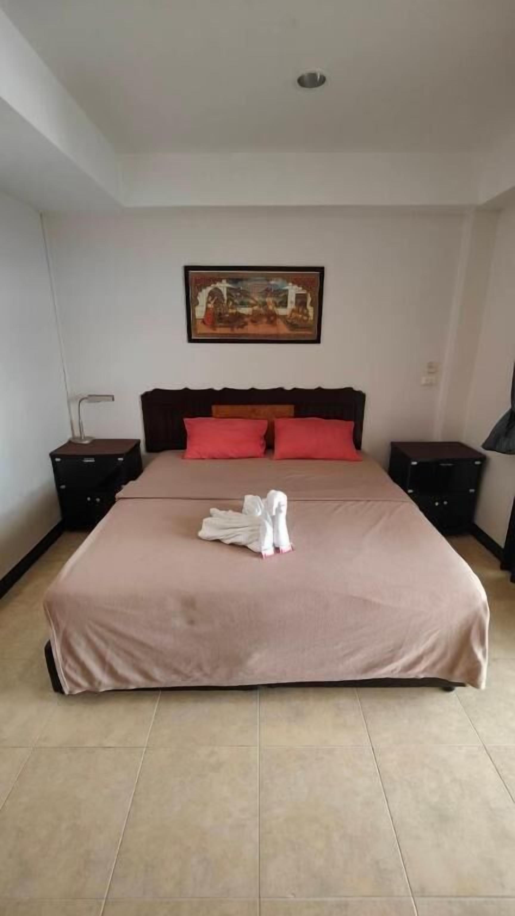 A neatly made bed with a pink bedspread and two red pillows, a painting on the wall, and two bedside tables with lamps.