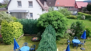 Un giardino curato con diversi arbusti e un alto conifero. Sono presenti diversi tavoli e sedie all'aperto, disposti per mangiare o rilassarsi.