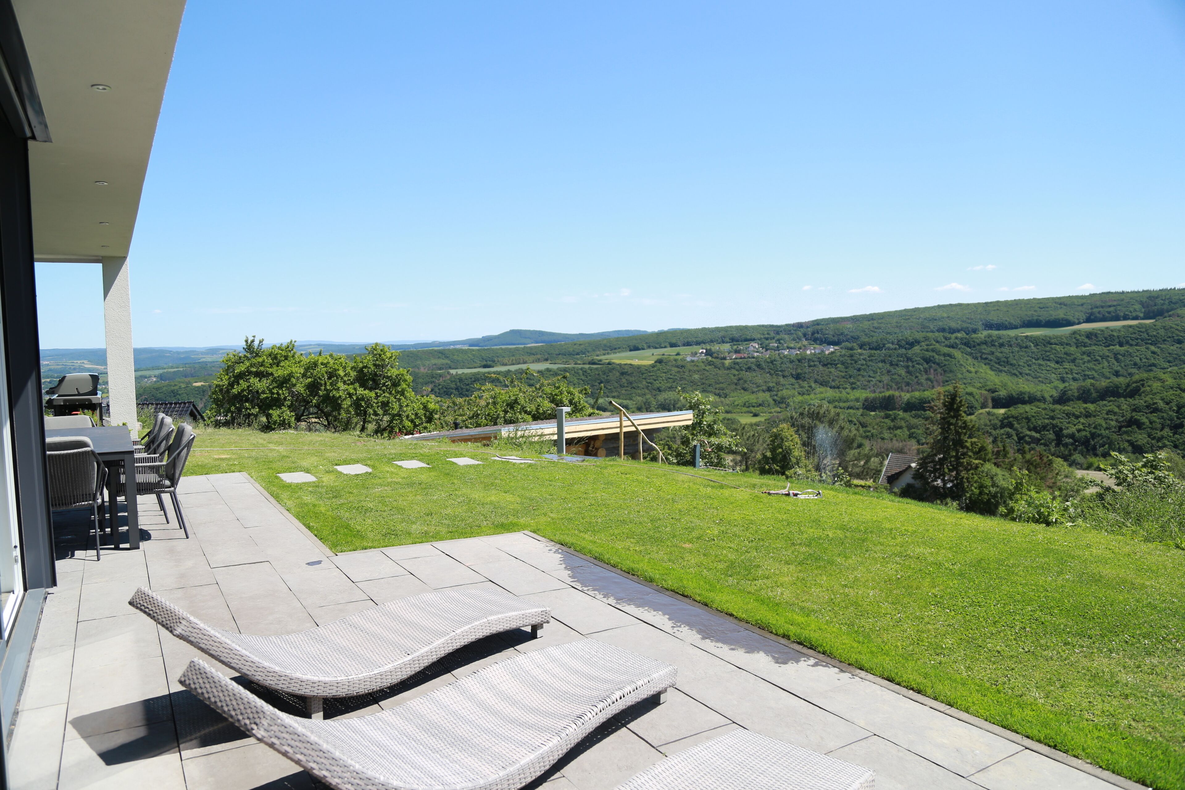 Un patio con due poltrone, un tavolo e sedie, affacciato su un paesaggio verdeggiante con colline e cielo sereno.