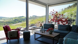 Un salon moderne doté d’une grande porte vitrée donnant sur une terrasse, d’un fauteuil rouge, d’un canapé bleu et offrant une vue sur des collines verdoyantes.