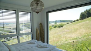 Une chambre à coucher avec un grand lit, du linge de lit blanc et une vue sur un paysage verdoyant par une grande fenêtre.