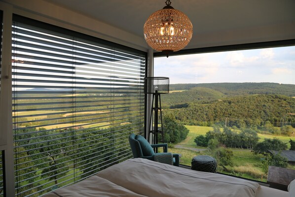 A bedroom with a large window, a bed, a chair, and a hanging light fixture.