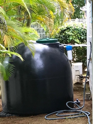 Ein großer, schwarzer Wassertank mit grüner Deckel, verbunden mit einem Schlauch, in einer Außenanlage mit weißem Zaun und Grünpflanzen im Hintergrund.