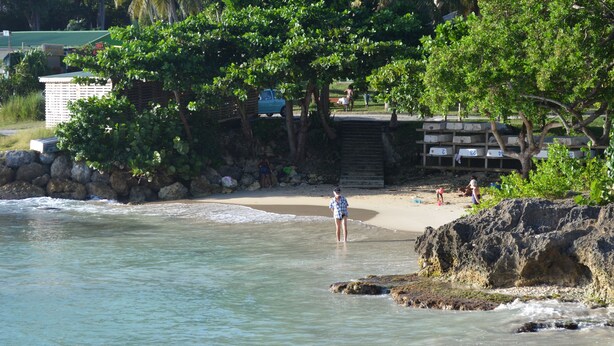 A beach with clear water, a rocky shoreline, and a few people. There are trees and a building in the background. - Cozy and modern charming villa 5 minutes walk from the beach (Le Gosier)