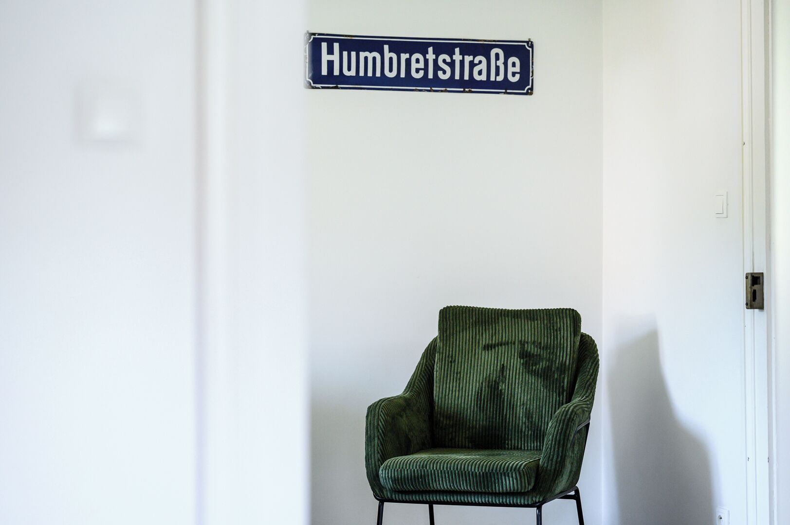 A green upholstered chair in a room with a white wall and a street sign reading 'Humbretstraße'.