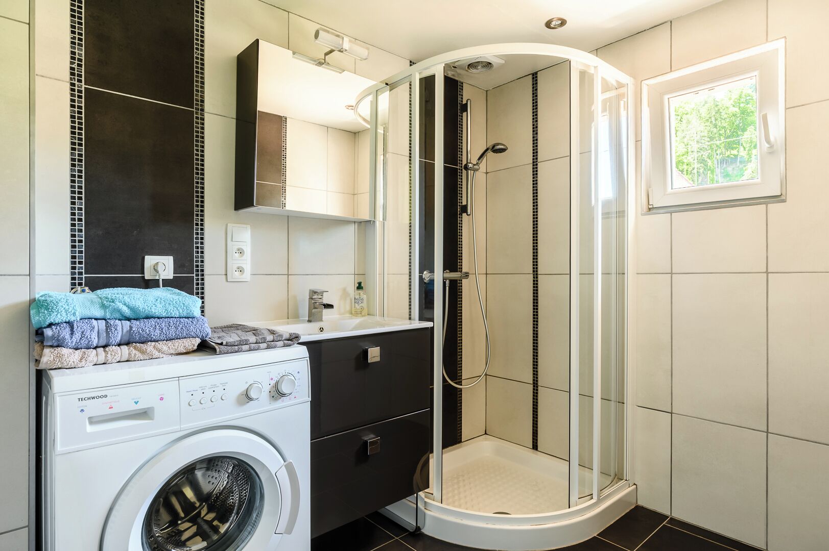 A modern bathroom with a washing machine, a shower enclosure, and a window.