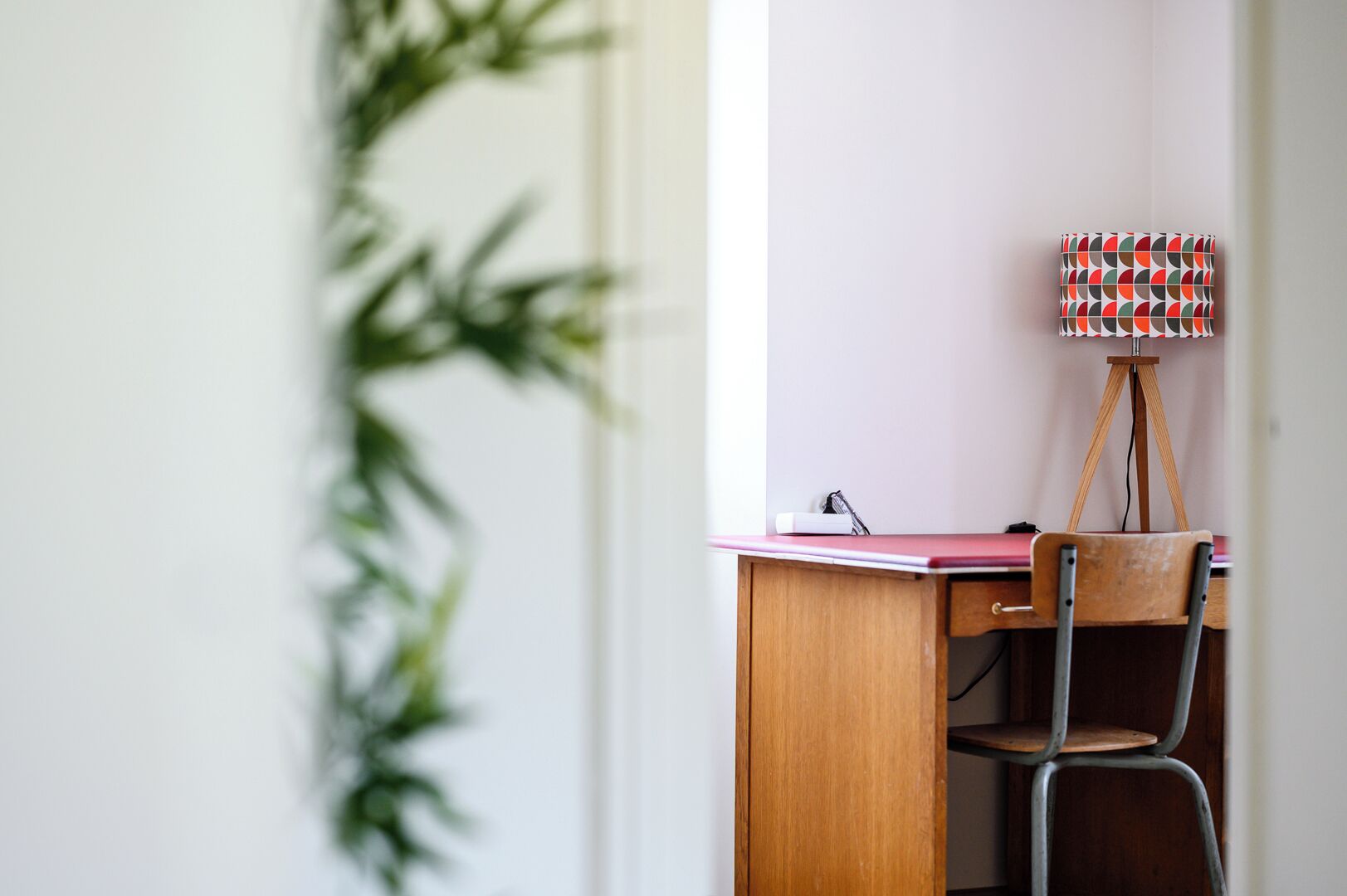 Un escritorio de madera con una silla, una lámpara y una planta en maceta en una habitación con pared blanca y una ventana.
