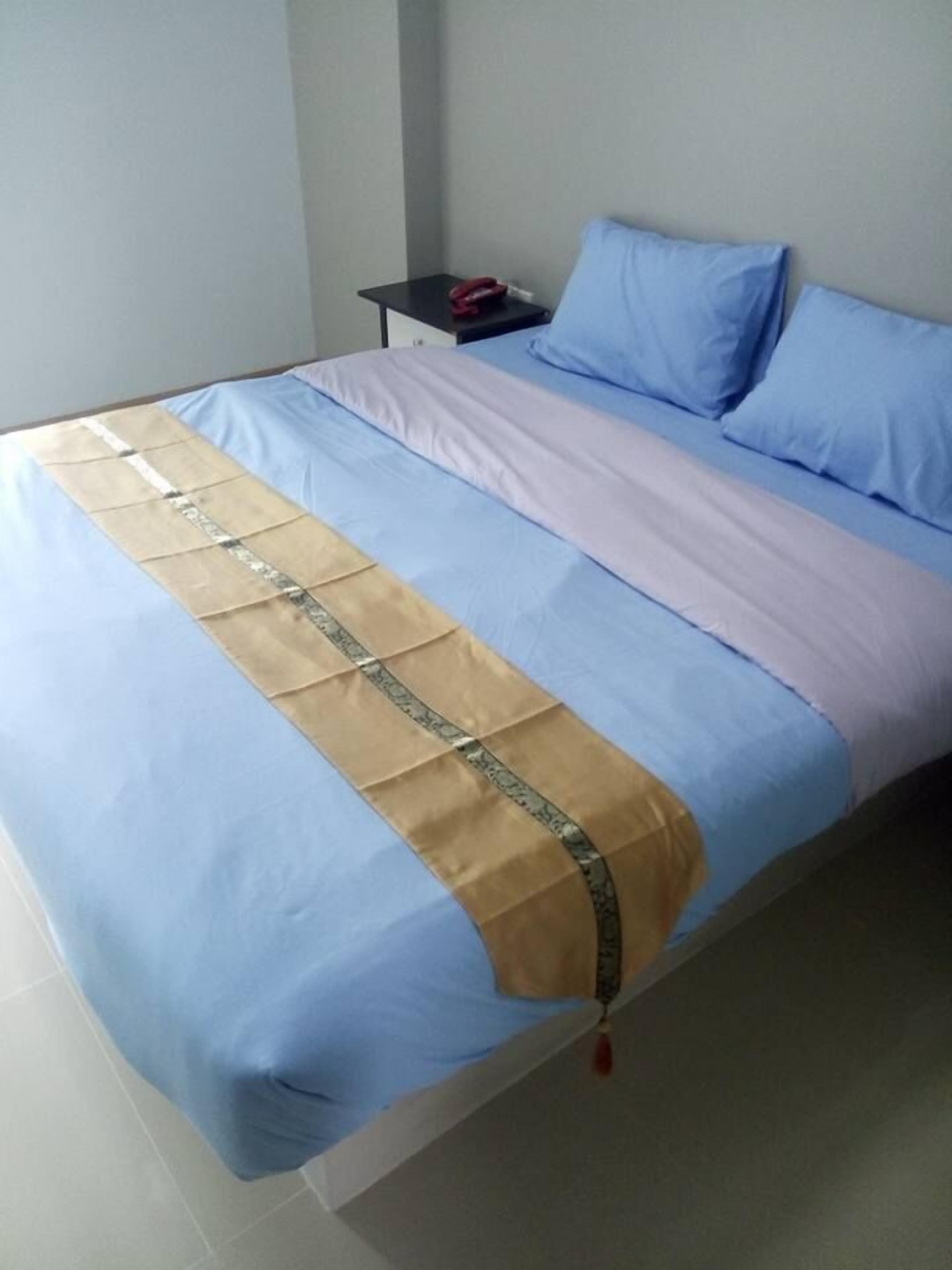A bed with a blue and beige bedspread, a small bedside table with a red object, and a light-colored wall.