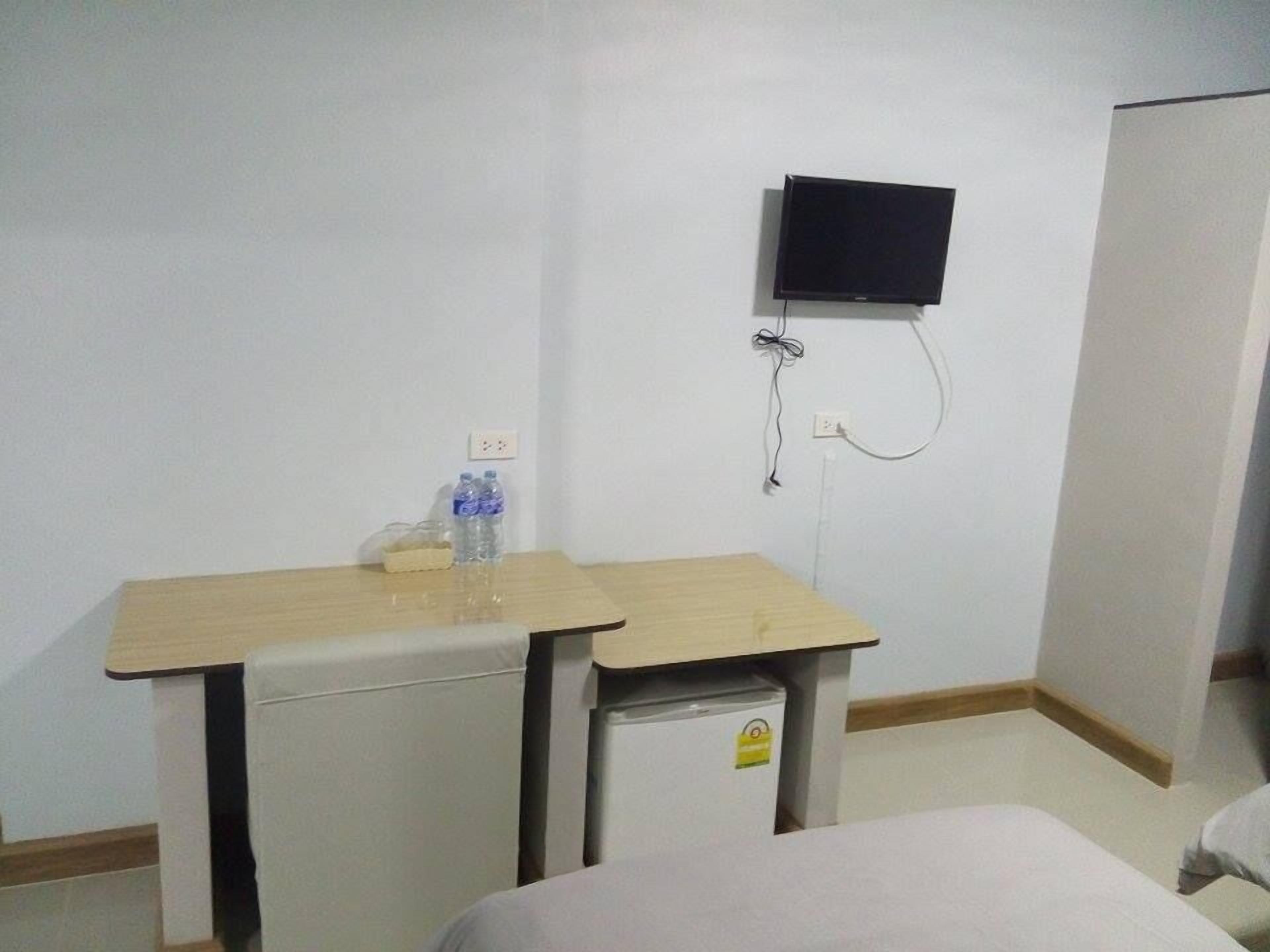 Ein kleines, schlichtes Zimmer mit einem Bett, einem Schreibtisch mit Stuhl, einem an der Wand montierten Fernseher und einem kleinen Kühlschrank.