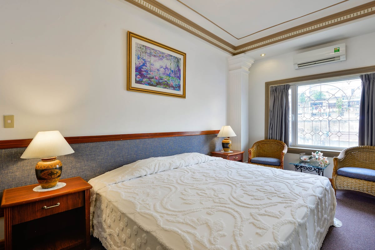 A hotel room with a large bed, two nightstands, a lamp, a framed painting, a window with curtains, and a small table with flowers.