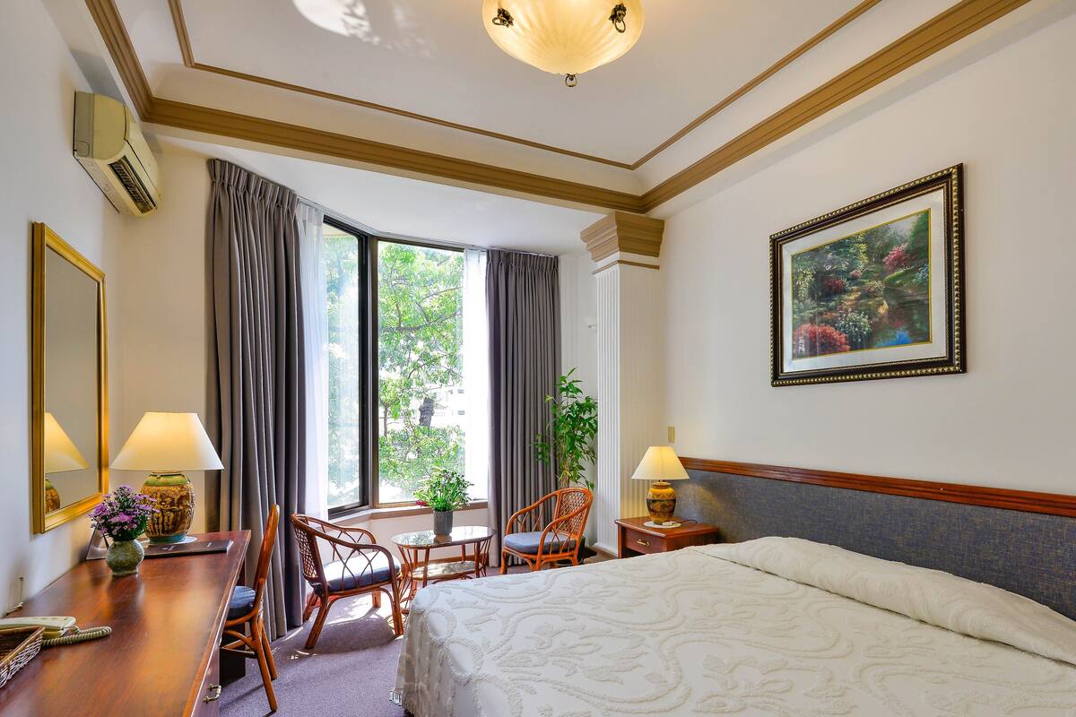 A hotel room with a bed, a desk with a lamp, a chair, a window with curtains, a plant, and a framed picture on the wall.