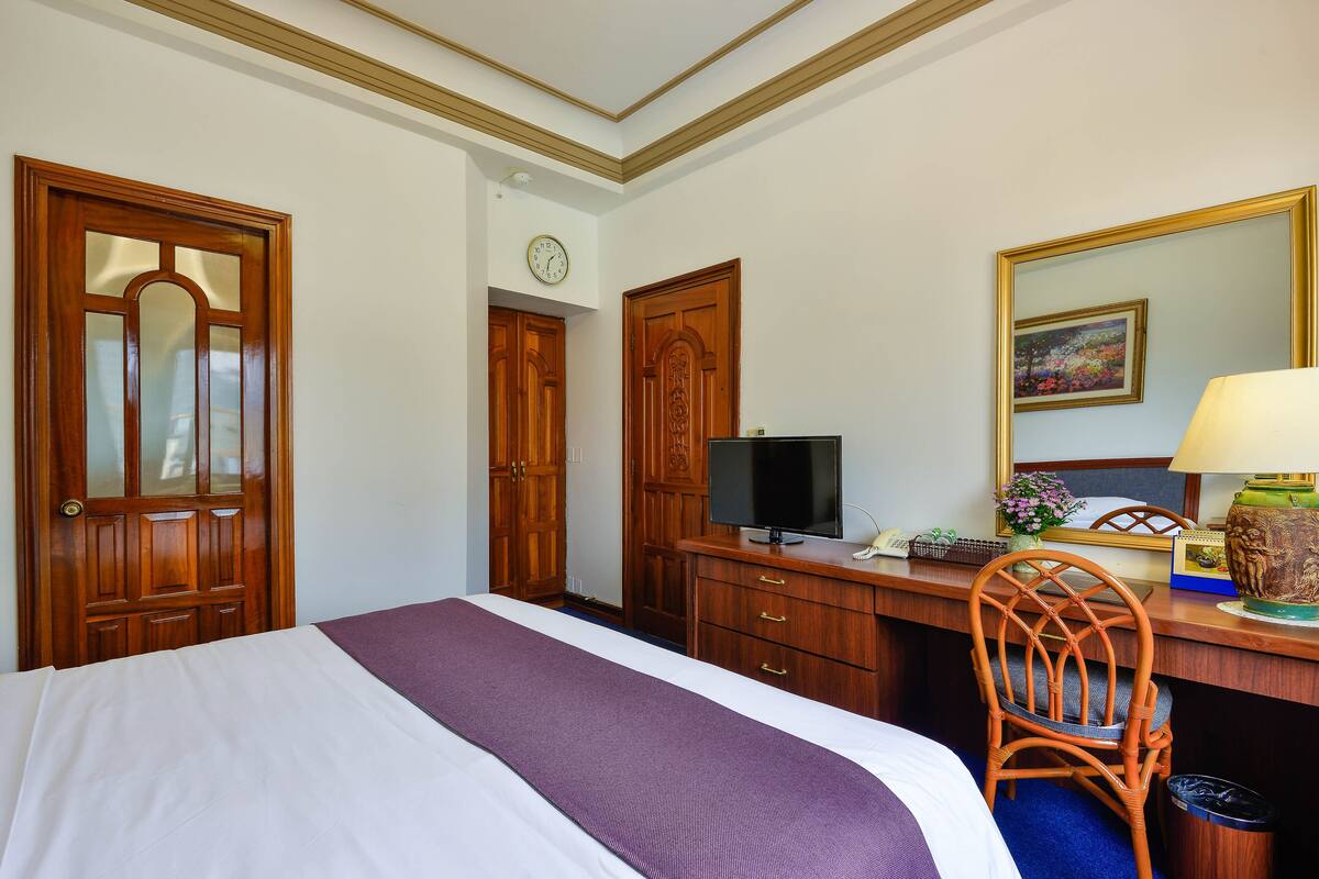 A hotel room with a bed, a desk with a computer, a chair, a desk lamp, and a trash can.