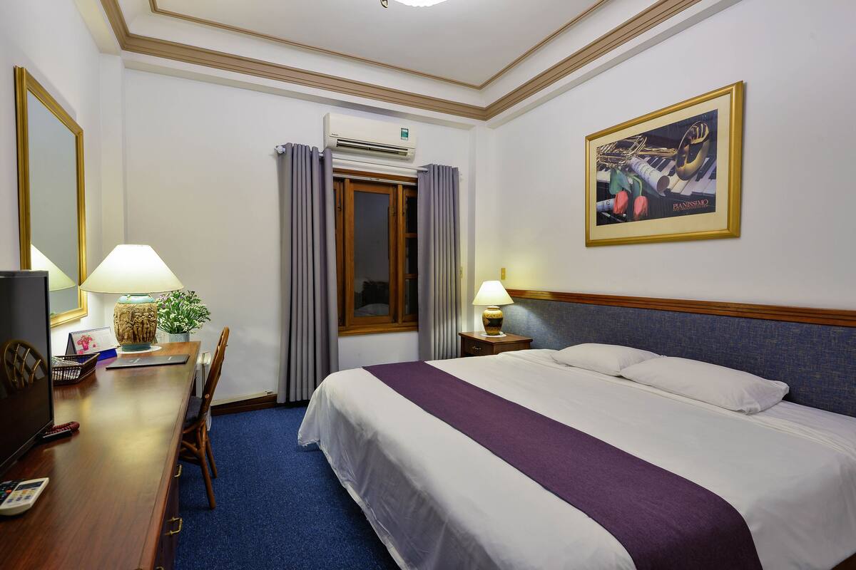 A hotel room with a bed, a desk with a lamp, a chair, a window with curtains, an air conditioning unit, and a framed picture on the wall.