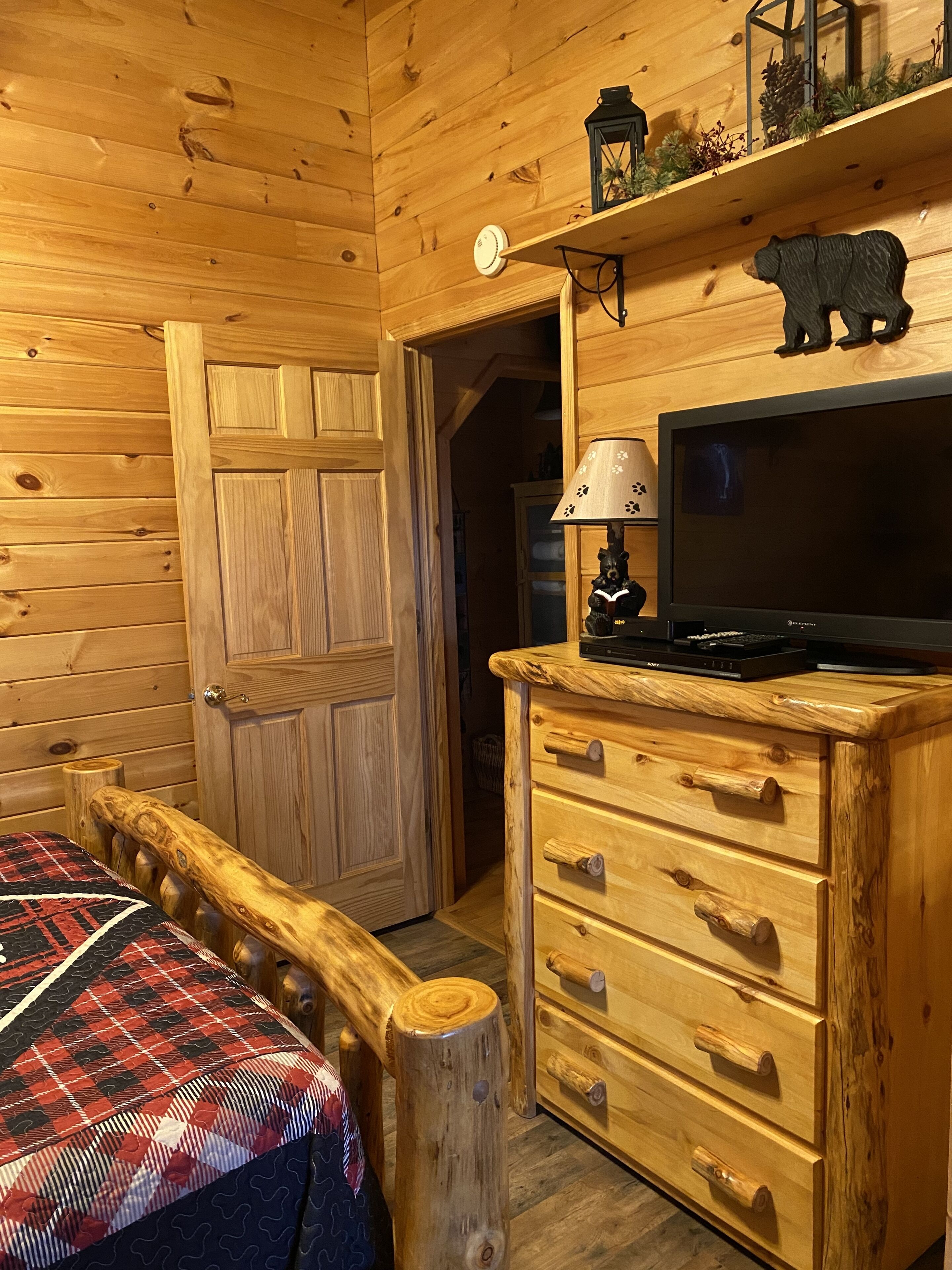 Een houten kamer met een bed, een dressoir, een televisie en een plank met decoratieve spullen.
