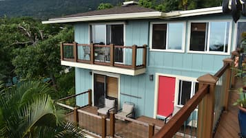 Une maison à deux étages avec un balcon, une porte rouge et une vue sur une colline boisée.