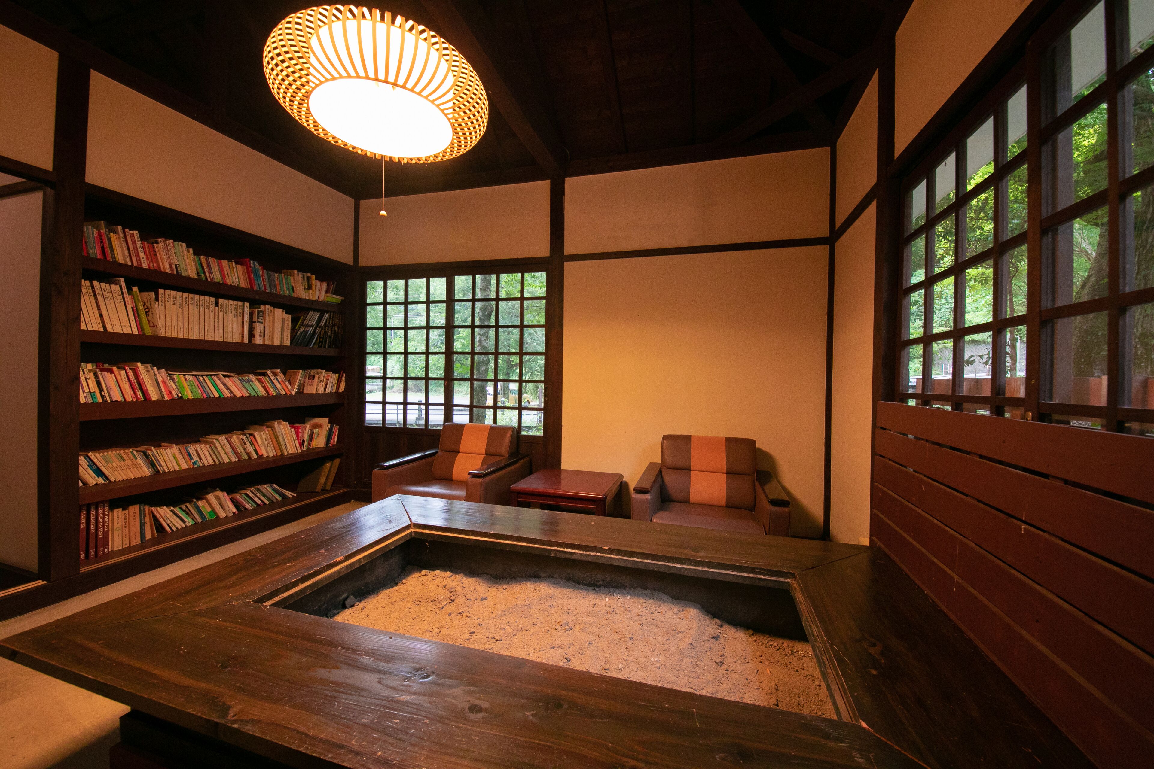 A traditional Japanese room with tatami flooring, a low wooden table, a bookshelf, and a large window with a view of greenery.