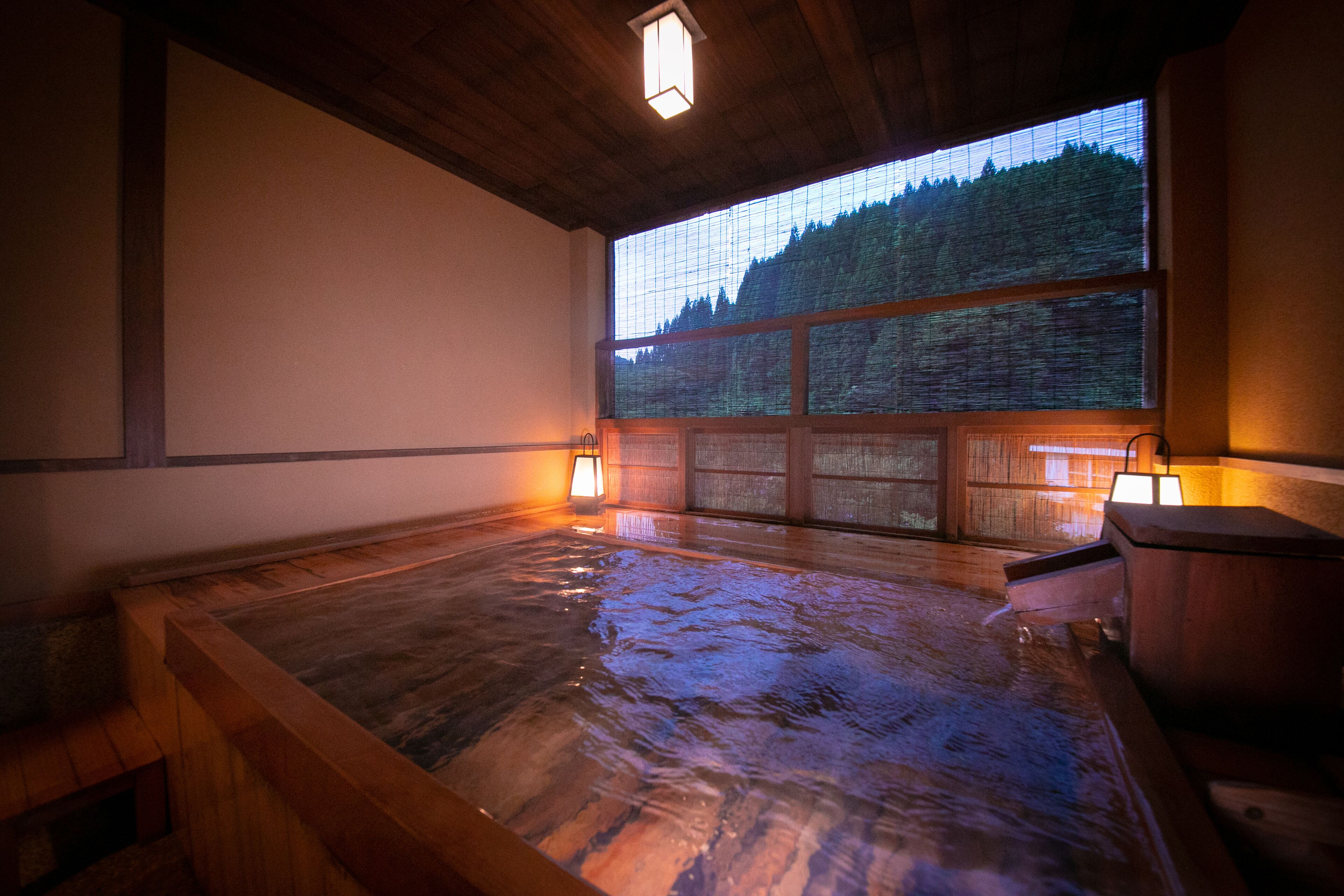 A traditional Japanese onsen with wooden flooring, a view of a forest through large windows, and two wall-mounted lanterns providing soft lighting.
