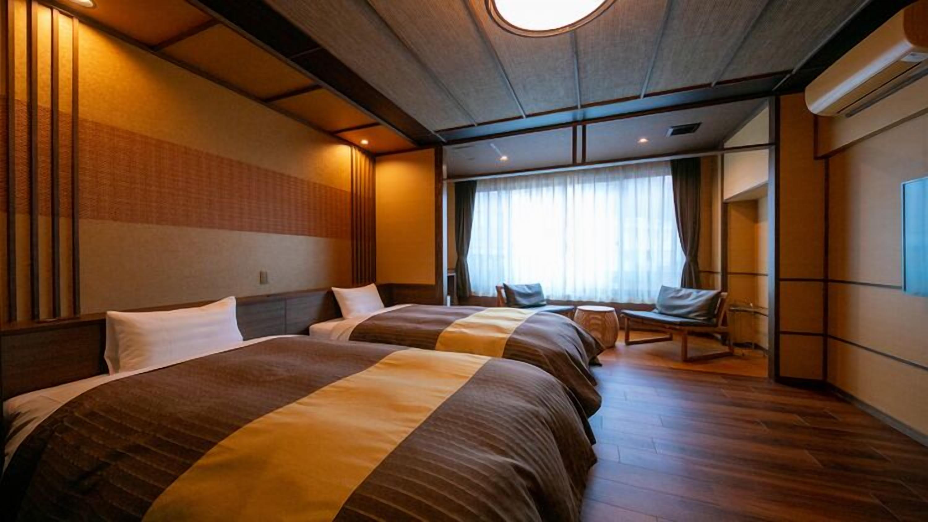 A hotel room with two beds, a sitting area with a chair, a large window with curtains, and a ceiling light.