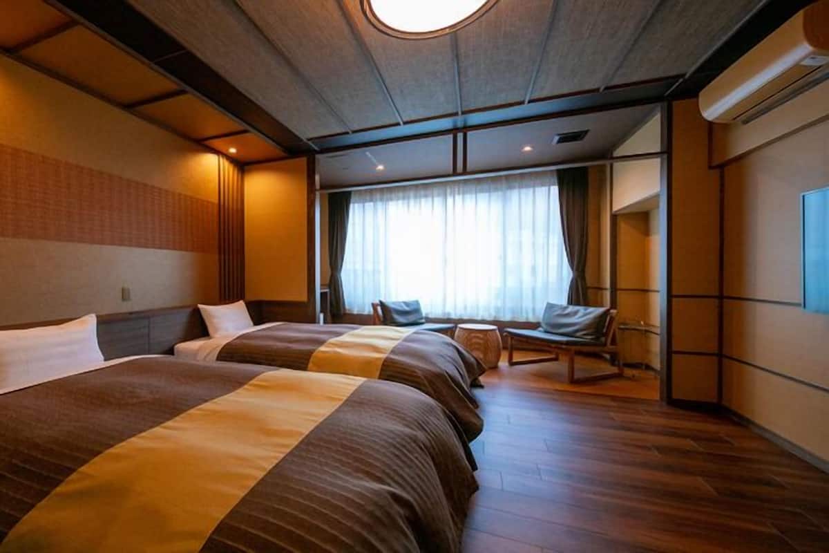 A hotel room with two beds, a sitting area with a chair, a large window with curtains, and a ceiling light.