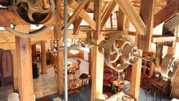 A rustic interior with exposed wooden beams, a dining area with wooden chairs, and a visible kitchen area. - River View. Rent Historic Mill's 2nd Floor. Rustic Timber-frame Construction (Shell Rock)