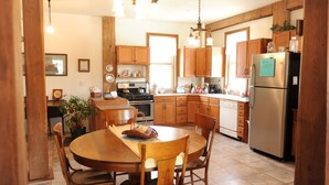 A kitchen with wooden cabinets, a stainless steel refrigerator, a dining table with chairs, and a hanging light fixture. - River View. Rent Historic Mill's 2nd Floor. Rustic Timber-frame Construction (Shell Rock)