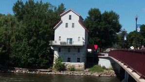 A two-story building with a red roof, located beside a river. - River View. Rent Historic Mill's 2nd Floor. Rustic Timber-frame Construction (Shell Rock)