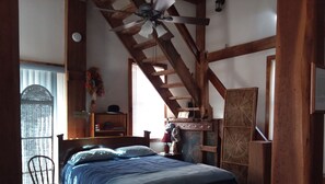 A studio apartment with a queen bed, a ceiling fan, a sliding glass door to the deck, and wooden beams. - River View. Rent Historic Mill's 2nd Floor. Rustic Timber-frame Construction (Shell Rock)