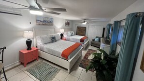 Quarto com duas camas, um ventilador de teto, mesa de cabeceira com luminária, uma cadeira e uma planta.