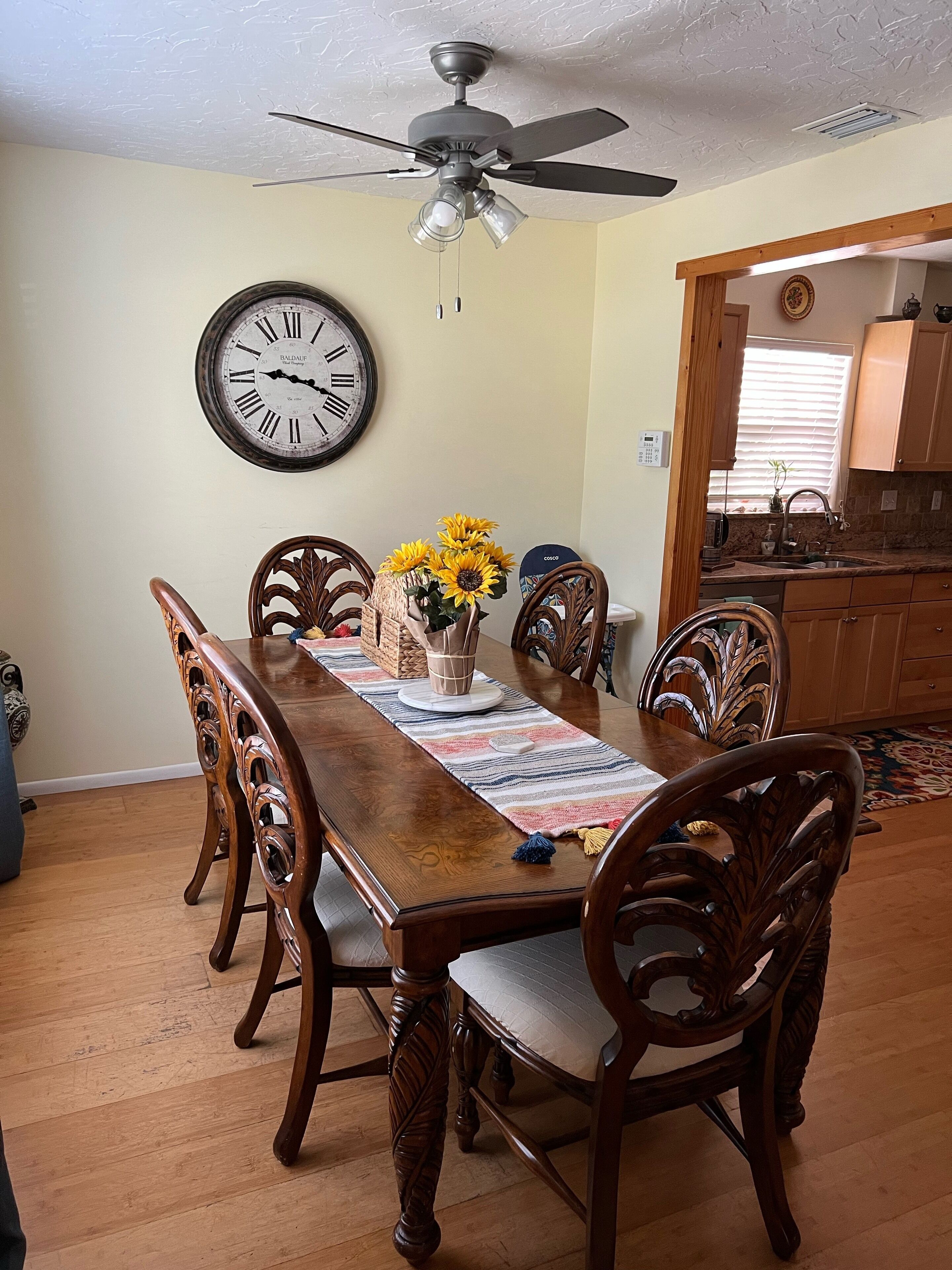 Comedor con mesa de madera, sillas, ventilador de techo, un reloj de pared y una cocina visible a través de un vano abierto.