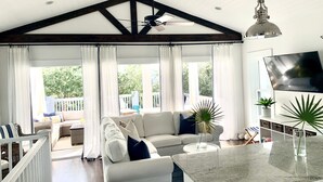 A spacious living area with a high ceiling, a ceiling fan, a large window with curtains, a white sofa, a coffee table, and a flat-screen TV mounted on the wall.