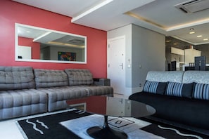 Uma sala de estar moderna com um sofá cinzento, um sofá preto com almofadas estampadas e uma mesa de centro de vidro, além de uma parede em destaque na cor vermelha.