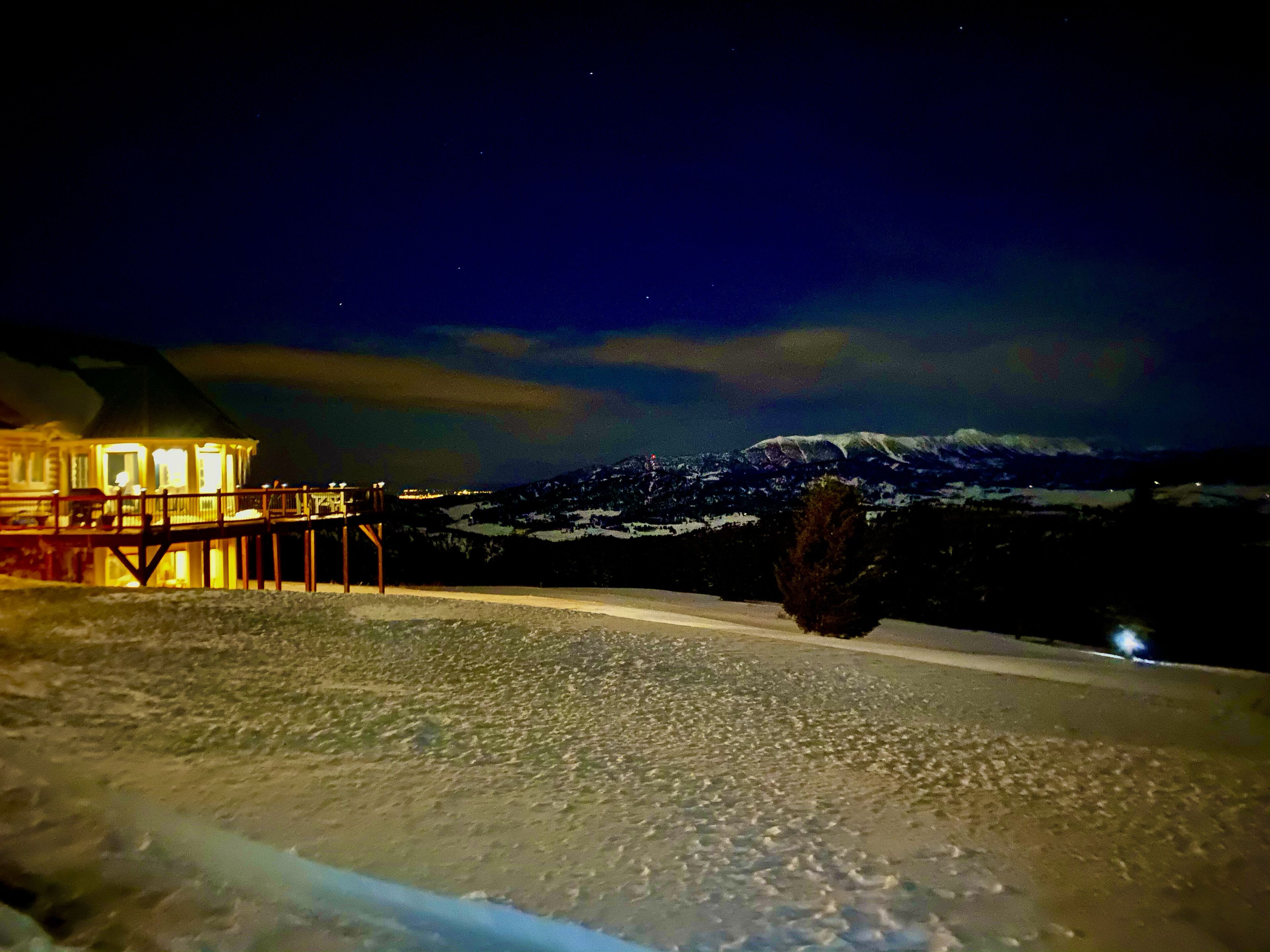 Paisaje nevado con una estructura de madera iluminada por luces cálidas, presumiblemente una cabaña o refugio, con vistas a una cordillera.